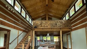 Un interior espacioso con alto techo, vigas de madera, una escalera central, comedor y vistas al exterior a través de amplias ventanas.