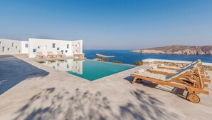 Et moderne hotel med pool og liggestole, med udsigt over et klart blåt hav og et klippefyldt landskab.
