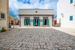 A cobblestone courtyard with a building featuring a central entrance, green shutters, and a small outdoor seating area.