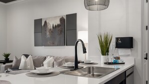 A modern kitchen with a white countertop, stainless steel sink, and a black faucet. There is a painting on the wall, a potted plant, and a Wi-Fi router on the counter. - 2 Bedroom, Main Level- ADA Accessible (Helena)