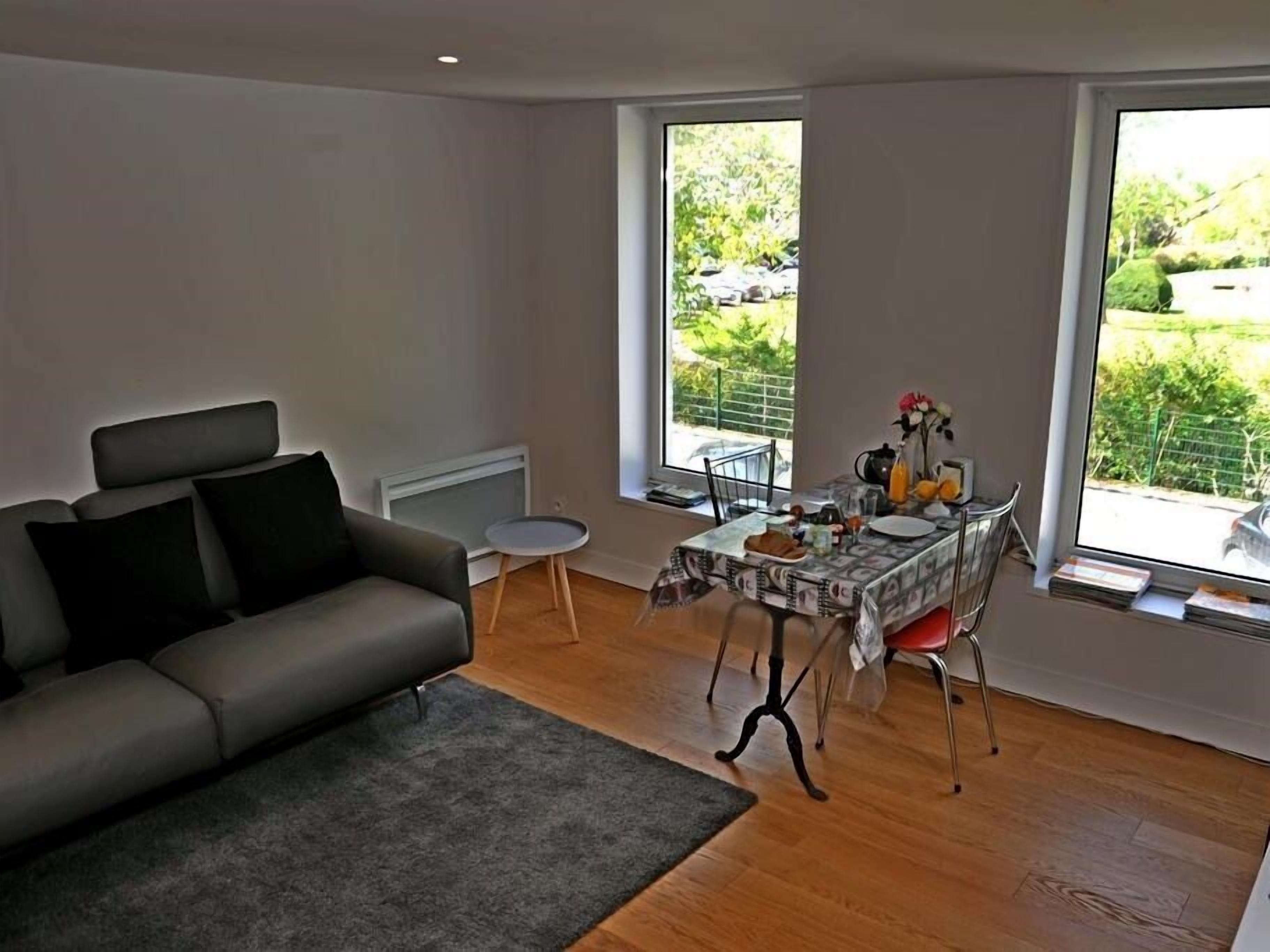 A modern living room with a grey sofa, a small round table, and a dining table with a view of the garden through large windows.