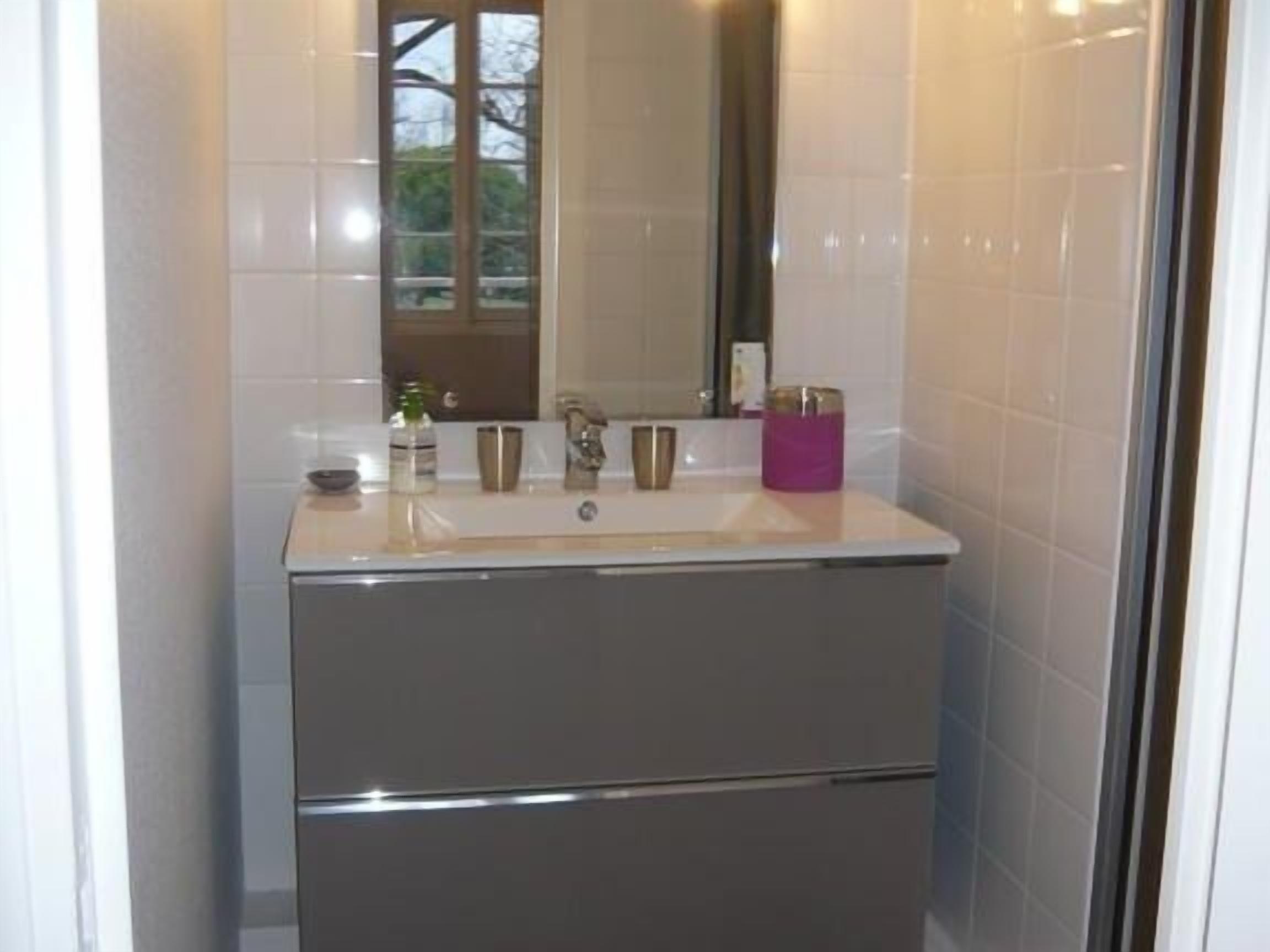 Un baño con lavamanos blanco, espejo y ventana con vista a árboles.
