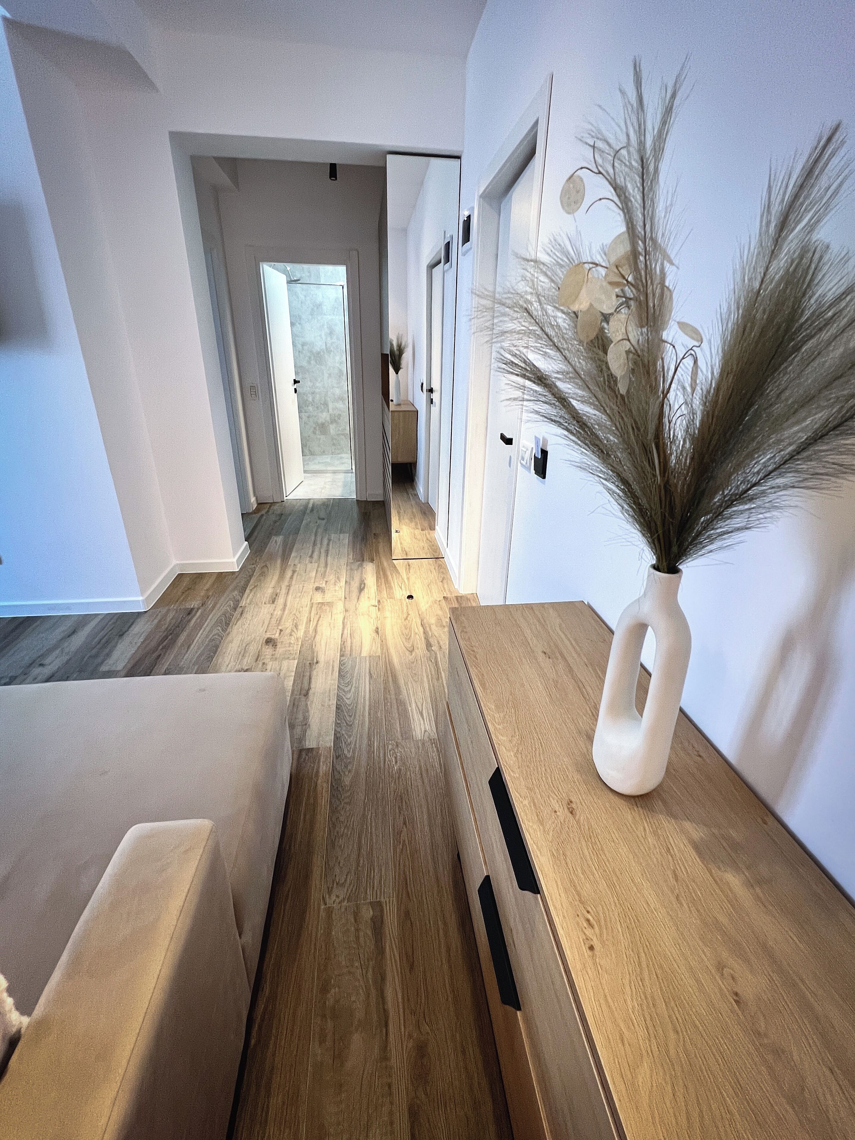Un couloir au sol en bois, un vase blanc contenant des plantes séchées et une porte donnant sur une autre pièce.