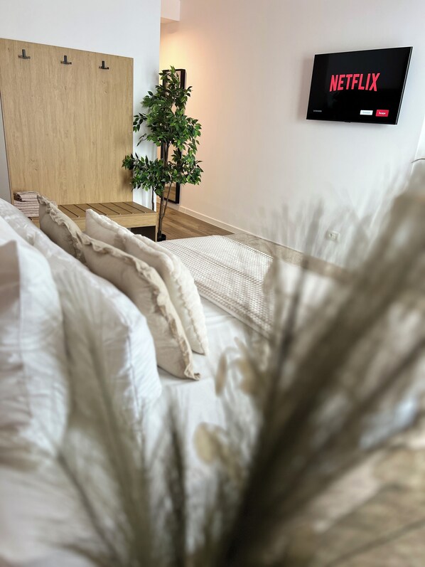 Un dormitorio con cama, almohadas, un armario de madera, una planta pequeña y un televisor de pared mostrando el logotipo de Netflix.