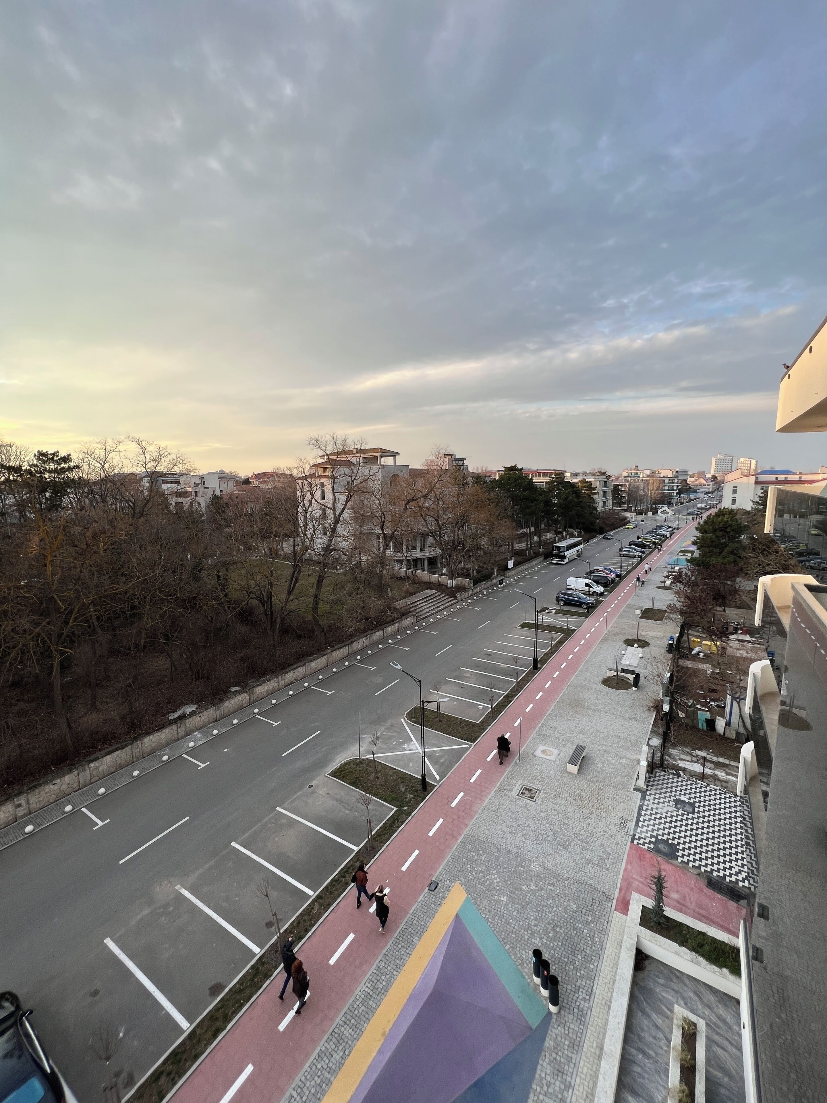 A city street with a pedestrian path, parked cars, and buildings in the background.