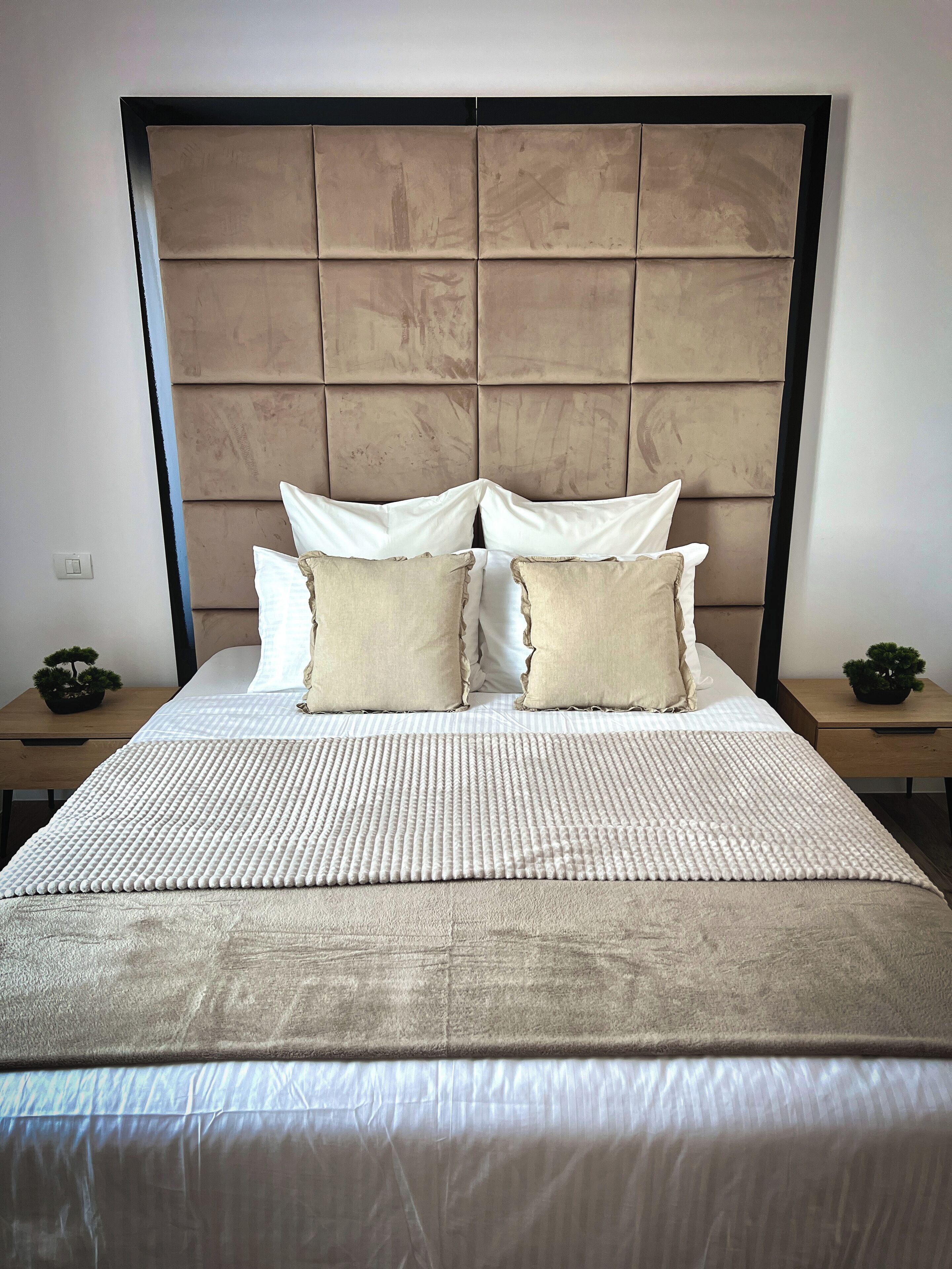 A neatly made bed with a beige headboard, white pillows, and a textured white bedspread. There are two bedside tables with small potted plants.