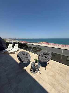 A terrace with two grey cushioned chairs, a small round table with a potted plant, and a glass balustrade overlooking a beach with red umbrellas. - Aparthotel Atmosphere (Eforie)
