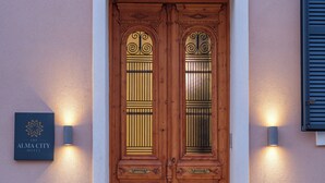 Une porte à double battant en bois, ornée de sculptures complexes, flanquée de deux lampes fixées au mur, et une enseigne au-dessus de la porte indiquant : « The Alma City Hotel ».