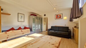 Ein modernes Wohnzimmer mit einer hölzernen Bank, einem grauen Sofa und einem Esstisch. Es gibt einen Kleiderschrank mit Glastüren, eine Uhr an der Wand und Kunstwerke.