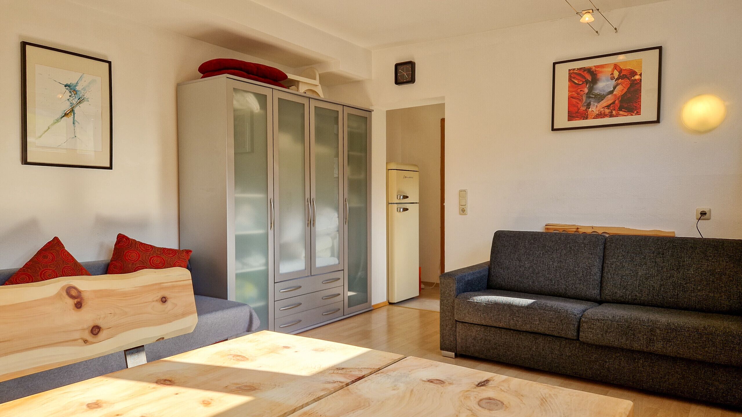 Una sala de estar con un sofá gris, una mesa de centro de madera, un aparador con puertas de cristal y dos cuadros en la pared.