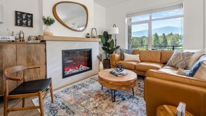 A living room with a fireplace, a sofa, a coffee table, and a view of the outdoors. - The Bauhaus | Private Hot Tub | Walk to Lake | MCM (Invermere)