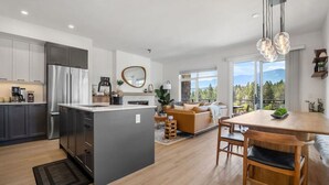 A modern kitchen with a central island, a dining area with a wooden table and chairs, and a living room with a sofa and a large window offering a view of the outdoors. - The Bauhaus | Private Hot Tub | Walk to Lake | MCM (Invermere)