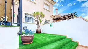 A well-maintained outdoor area with artificial green grass, potted plants, and a white wall with a 'NO' sign.