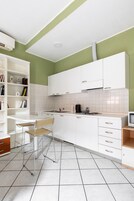 Una cocina moderna con armarios blancos, una estantería empotrada, un comedor con una silla y un televisor pequeño.