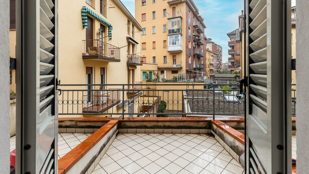 En balkon med fliset gulv, gelænder i metal og udsigt til omkringliggende bygninger.