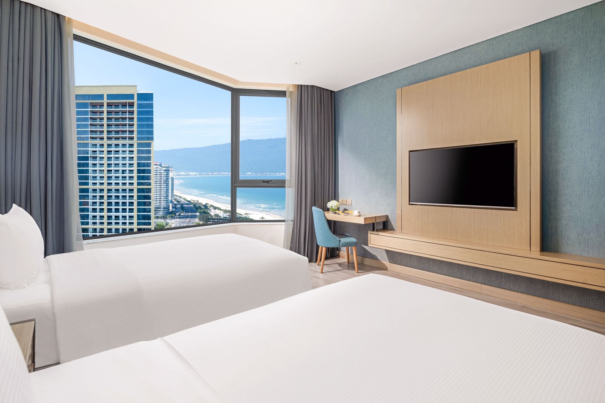 Habitación de hotel con dos camas, un escritorio, un televisor y un amplio ventanal con vistas a la ciudad.