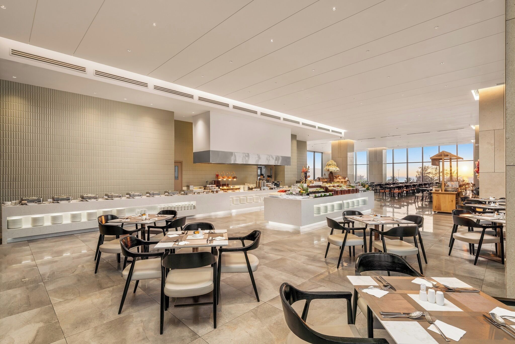 A spacious hotel dining area with multiple tables and chairs, a central buffet, and a large window offering a view.