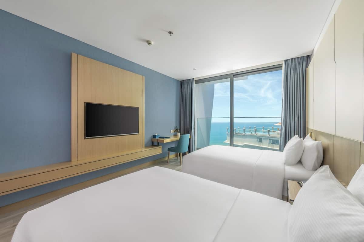 A hotel room with two beds, a TV mounted on the wall, a desk with a chair, and a large window offering a view of the sea.