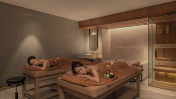 A spa room with two massage tables, a wooden ceiling, a glass sauna door, and a sink.