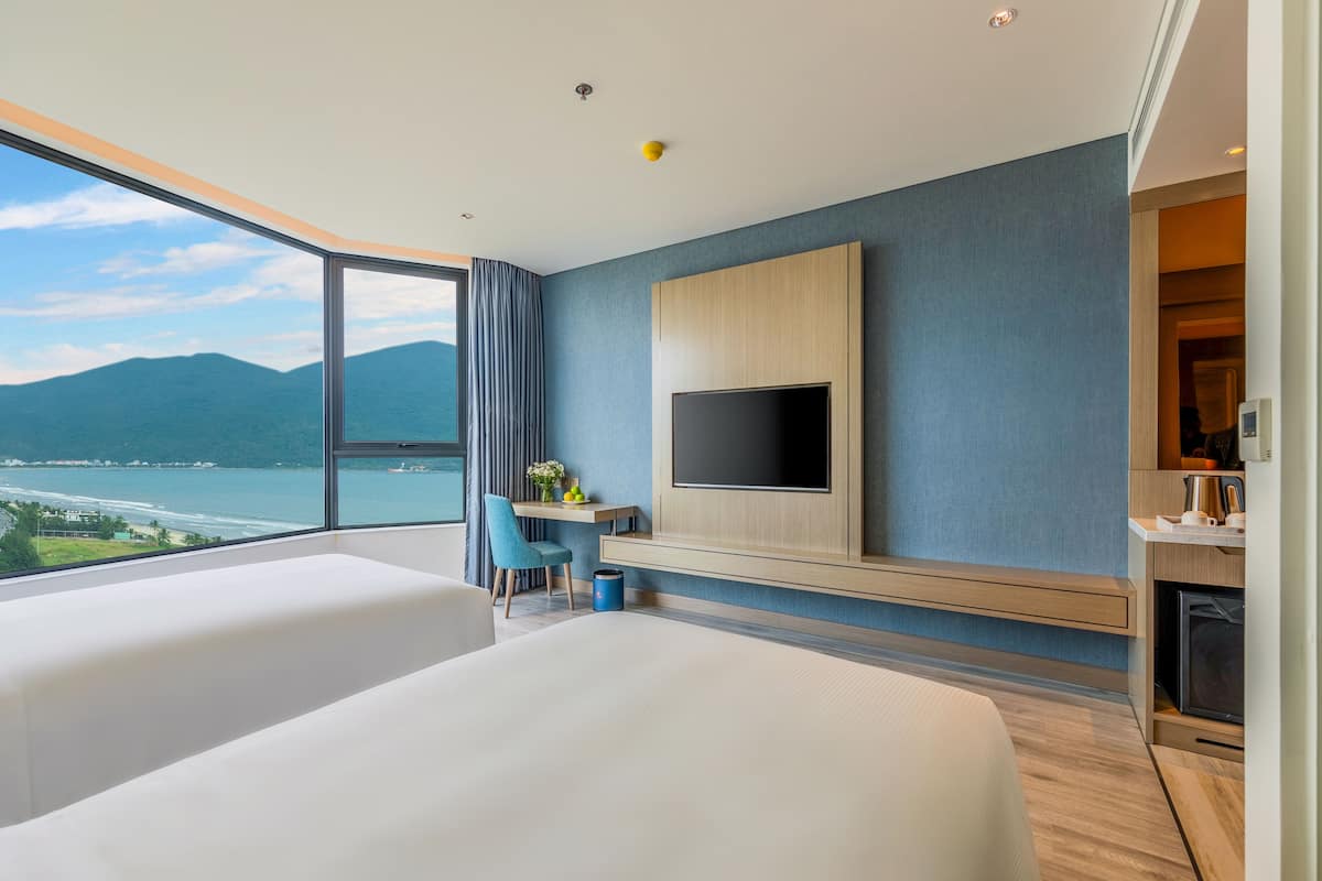 A modern hotel room with two beds, a TV, a desk, and a view of the sea.