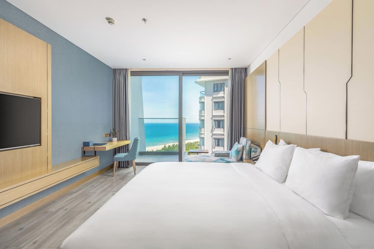A hotel room with a large bed, a desk, a chair, and a TV. The room has a view of the ocean and a building.