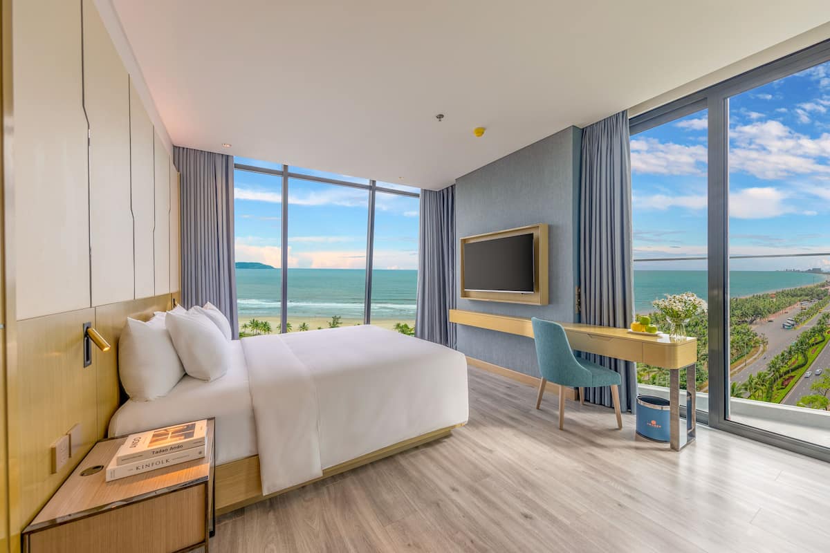 A modern hotel room with a large bed, a desk, a chair, a TV, and a view of the ocean.