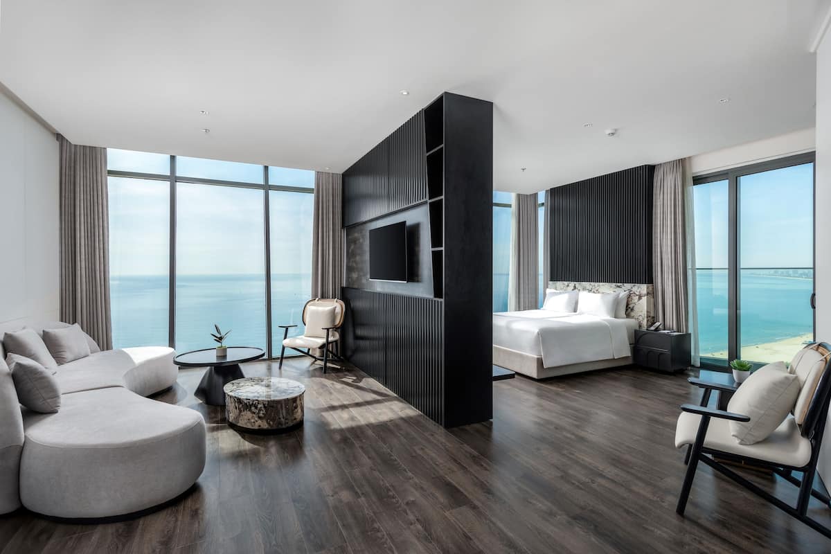 A modern hotel room with a large bed, a sofa, a TV, and a view of the ocean.