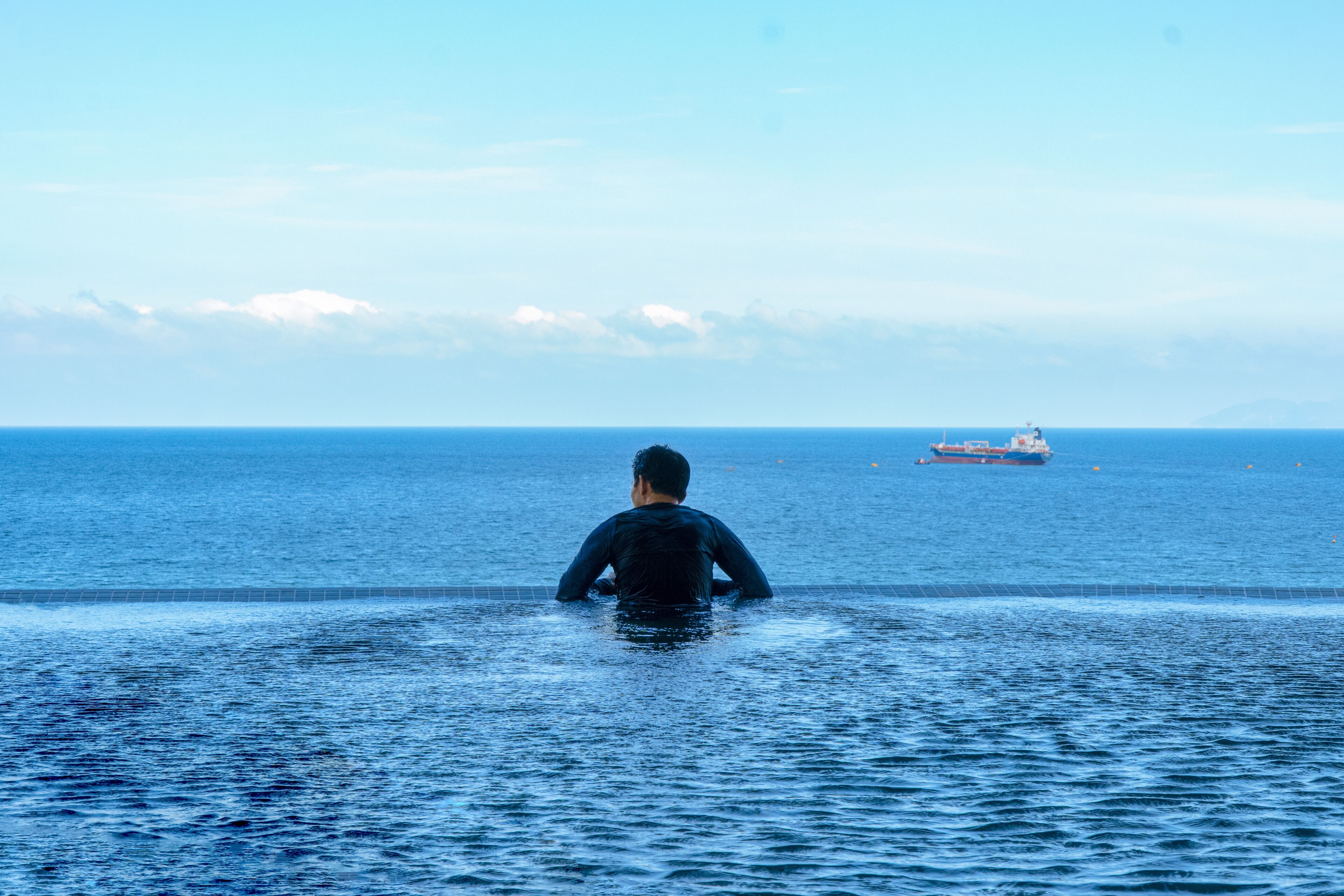 A person in a wetsuit standing in the water facing a ship.