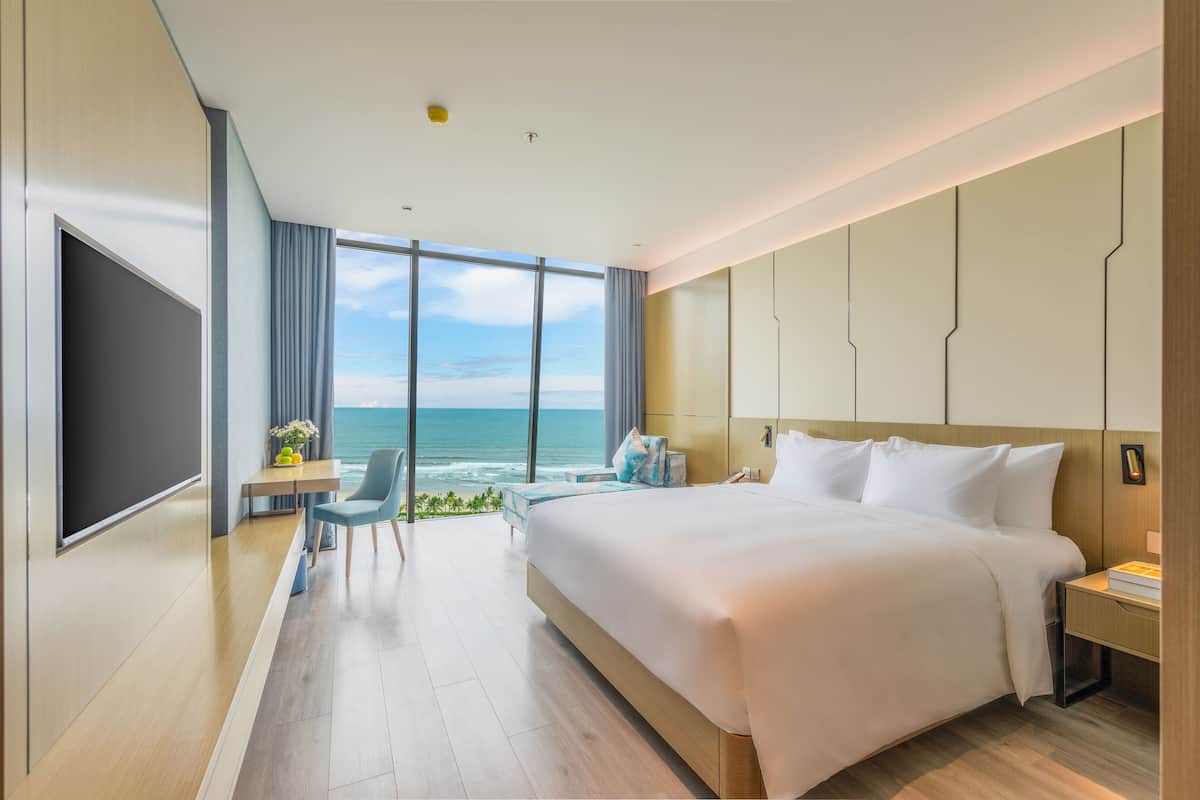 A modern hotel room with a large bed, a desk, and a view of the ocean.