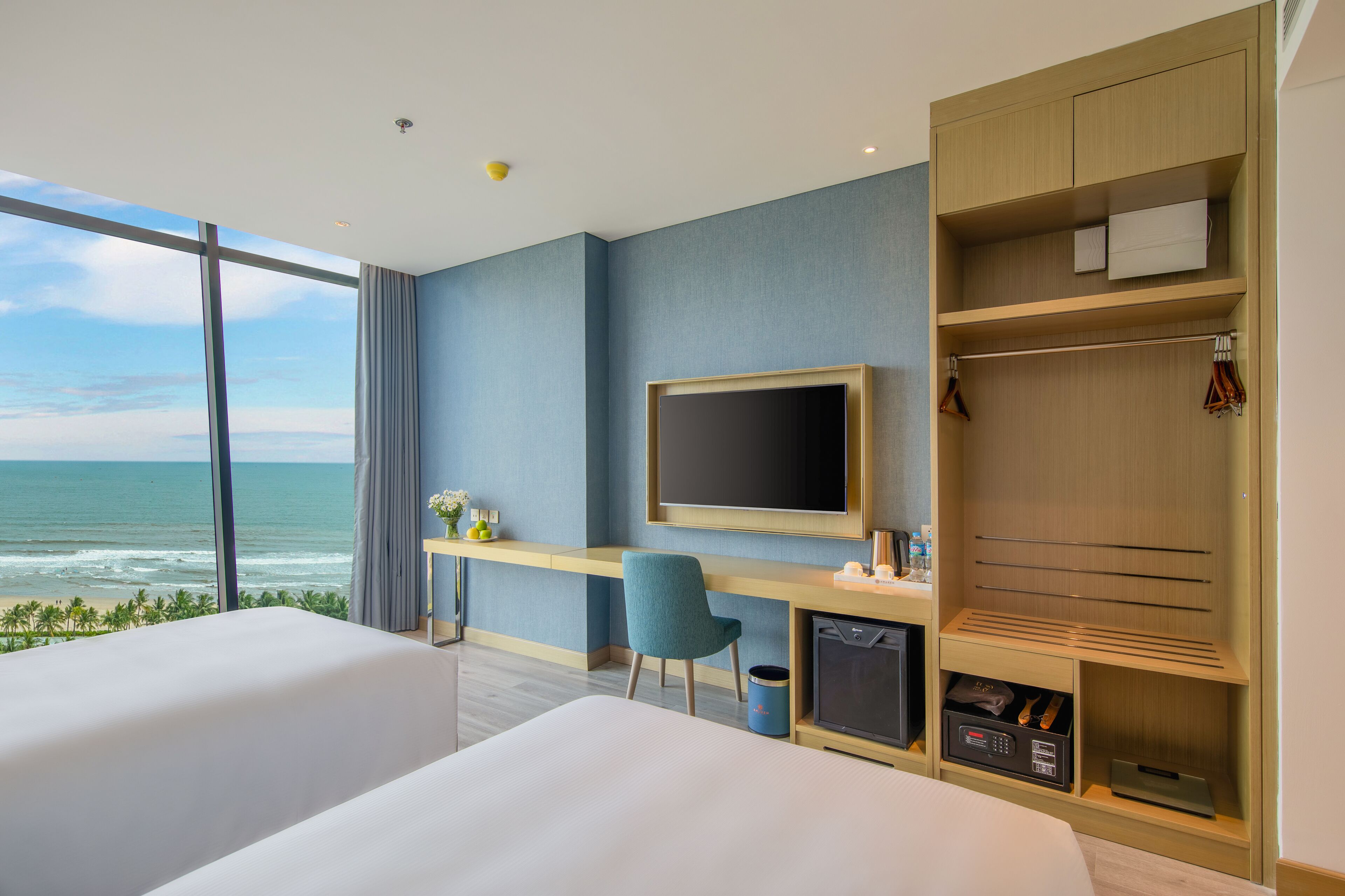 A hotel room with a large window overlooking the ocean, a desk with a chair, a TV, and a wardrobe.