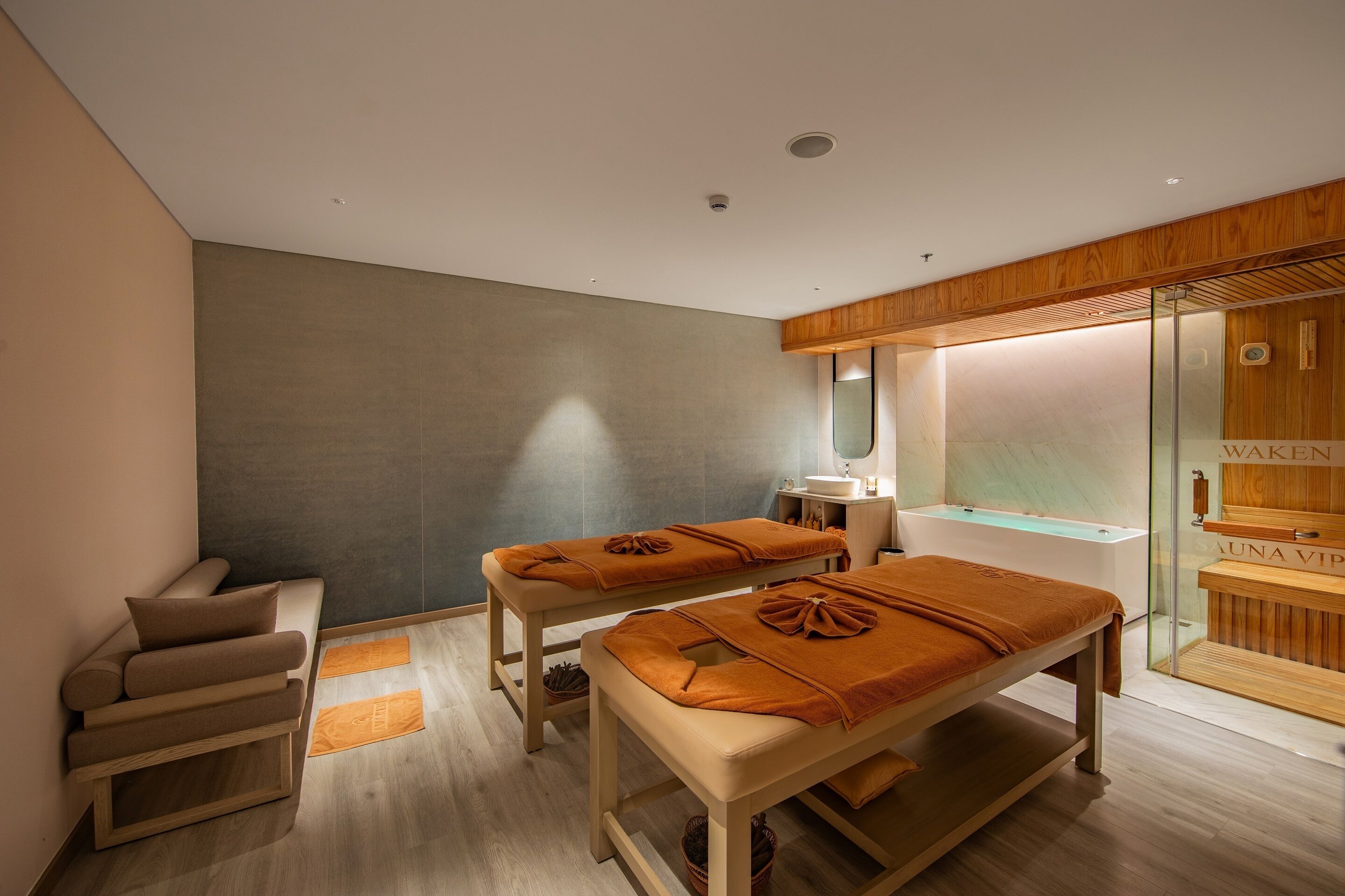 Une salle de spa équipée de deux tables de massage, d’un banc en bois, d’un espace de douche en verre et d’un comptoir d’accueil en bois.