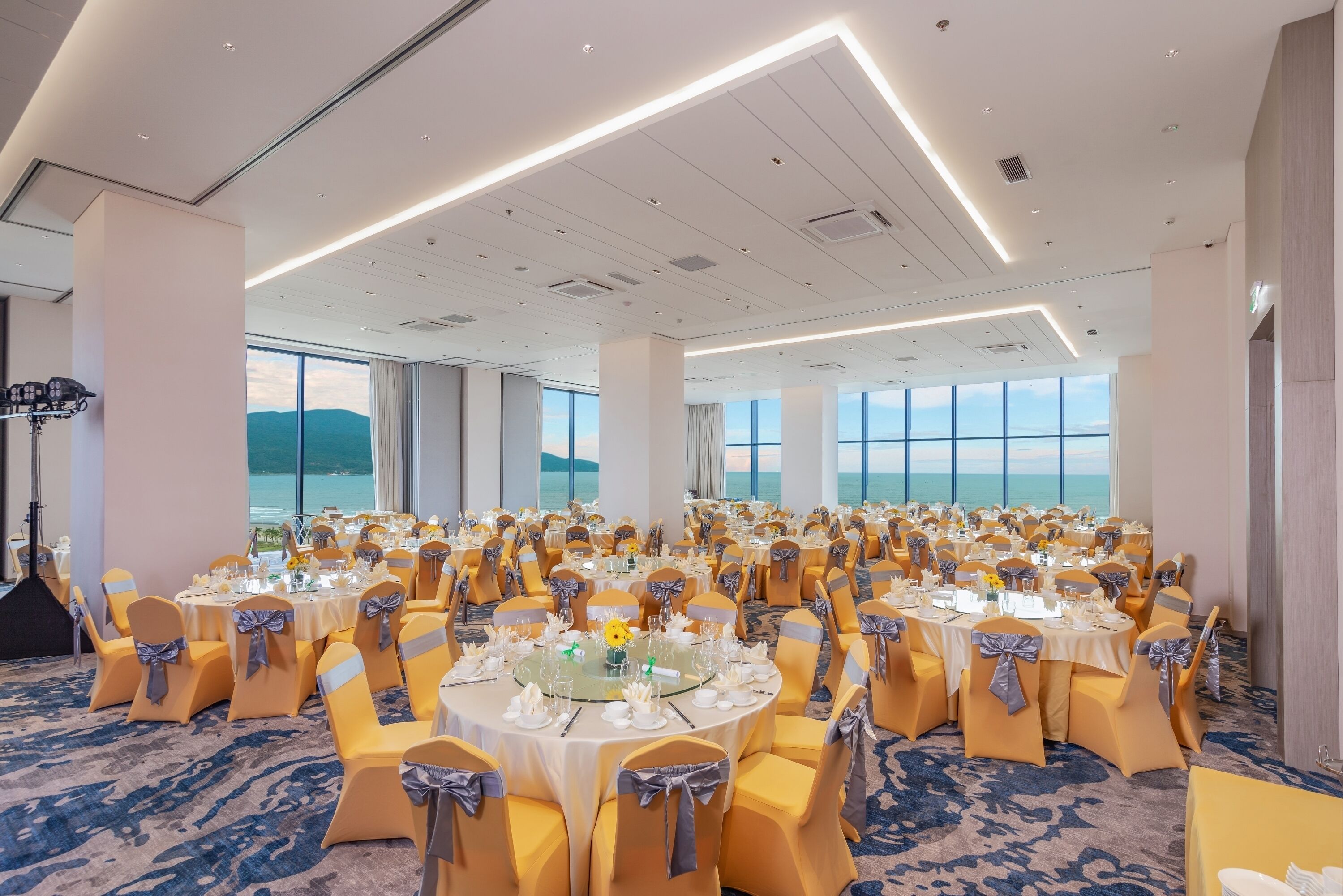 A large banquet hall with round tables set for a formal event, featuring a carpeted floor and large windows offering a view of the sea.