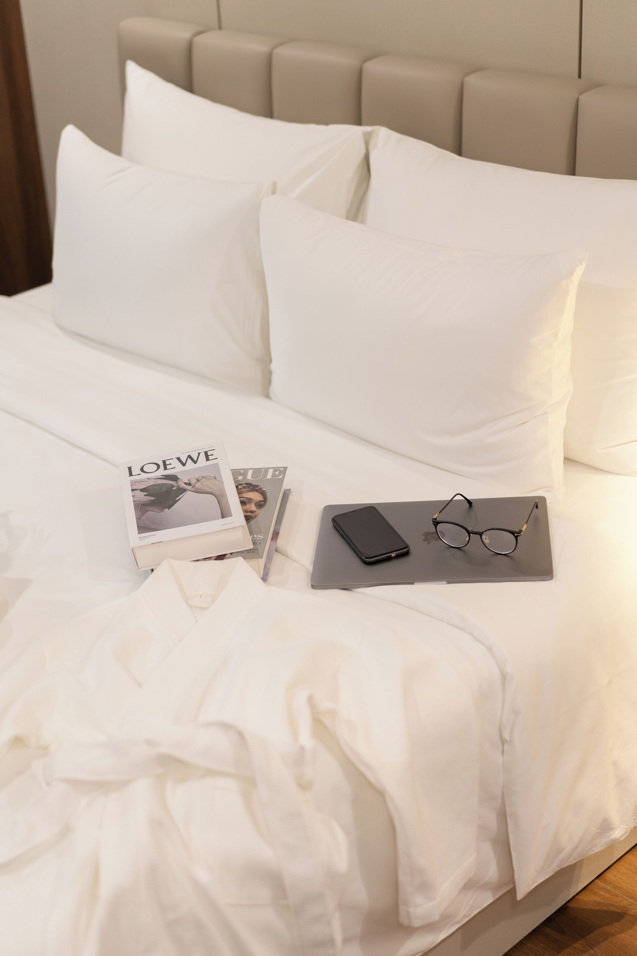 Una cama bien hecha con sábanas blancas, un par de gafas, un teléfono inteligente y una revista sobre una mesita de noche.