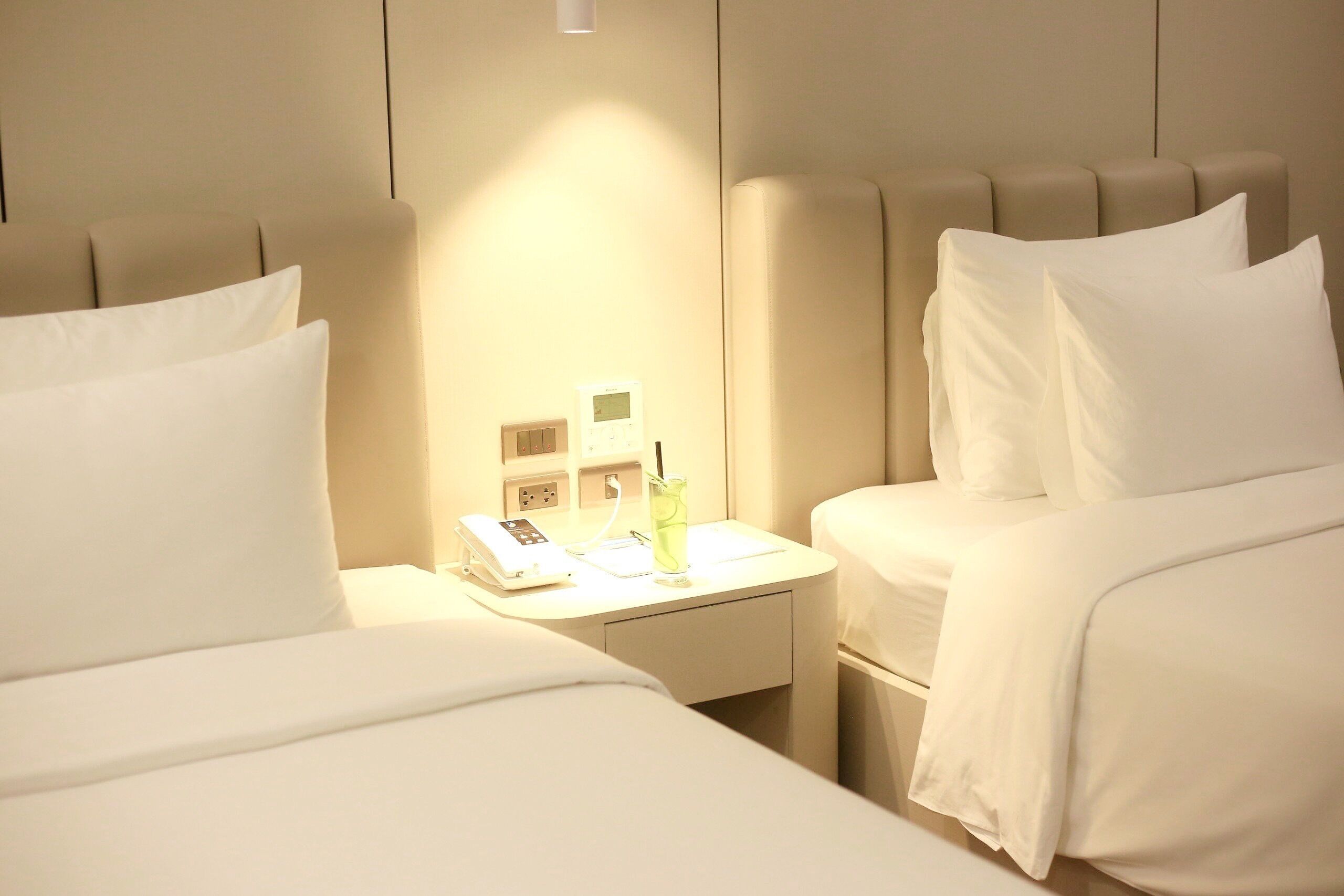 A hotel room with two beds, a bedside table with a phone and a glass of water, and a wall-mounted thermostat.