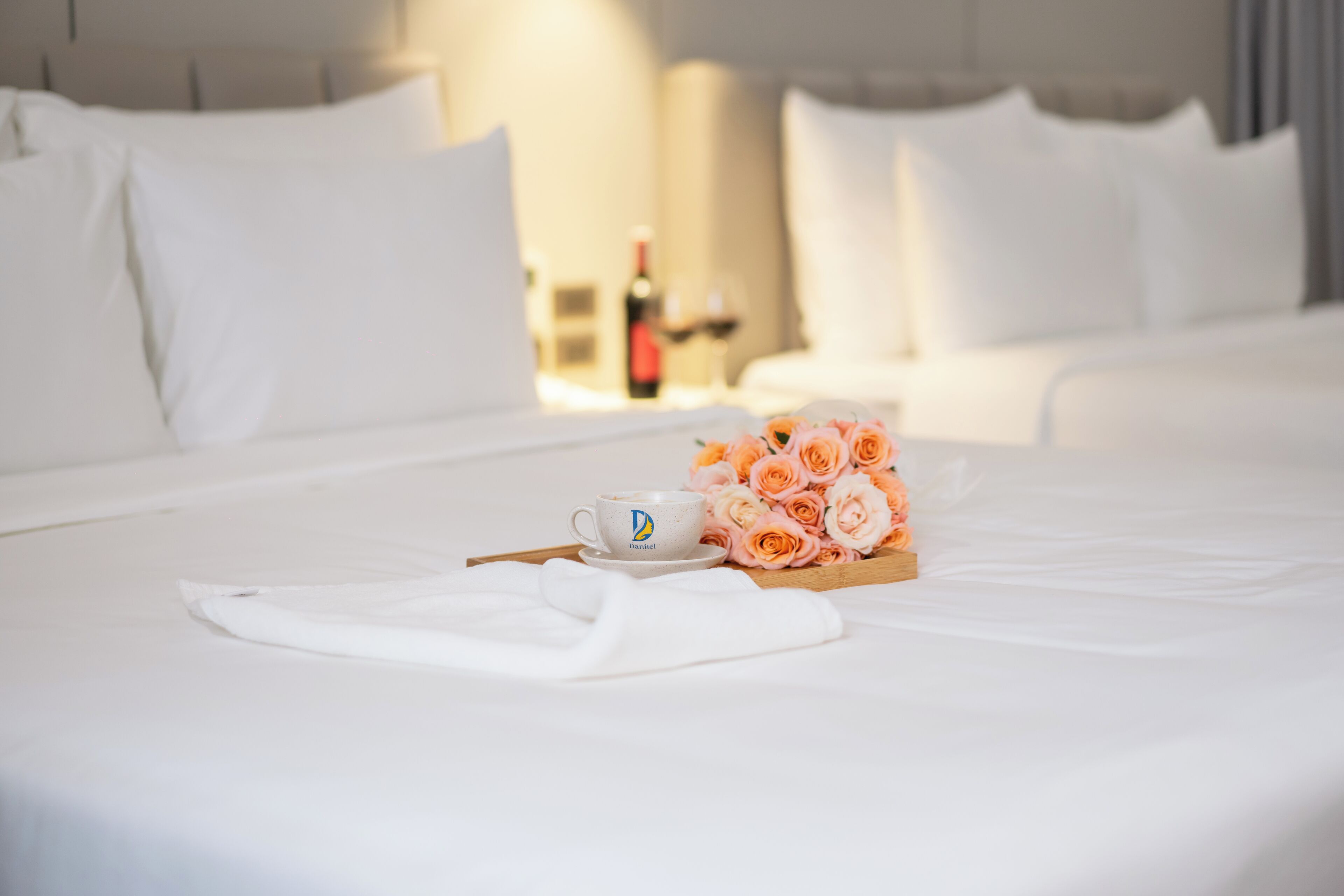 A neatly made bed with white linens, a cup with a logo, and a bouquet of roses on a wooden tray.