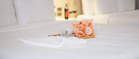 A neatly made bed with white linens, a cup with a logo, and a bouquet of roses on a wooden tray.
