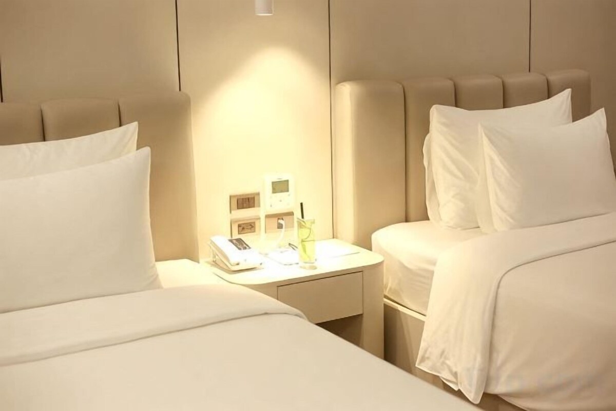 A hotel room with two beds, a bedside table with a phone and a bottle, and a wall-mounted light.