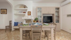 A dining area with a wooden table, chairs, and a fruit bowl. There is a TV on a stand and a shelf with decorative items. - Affaccio Calipso - PT by BarbarHouse (Marittima)