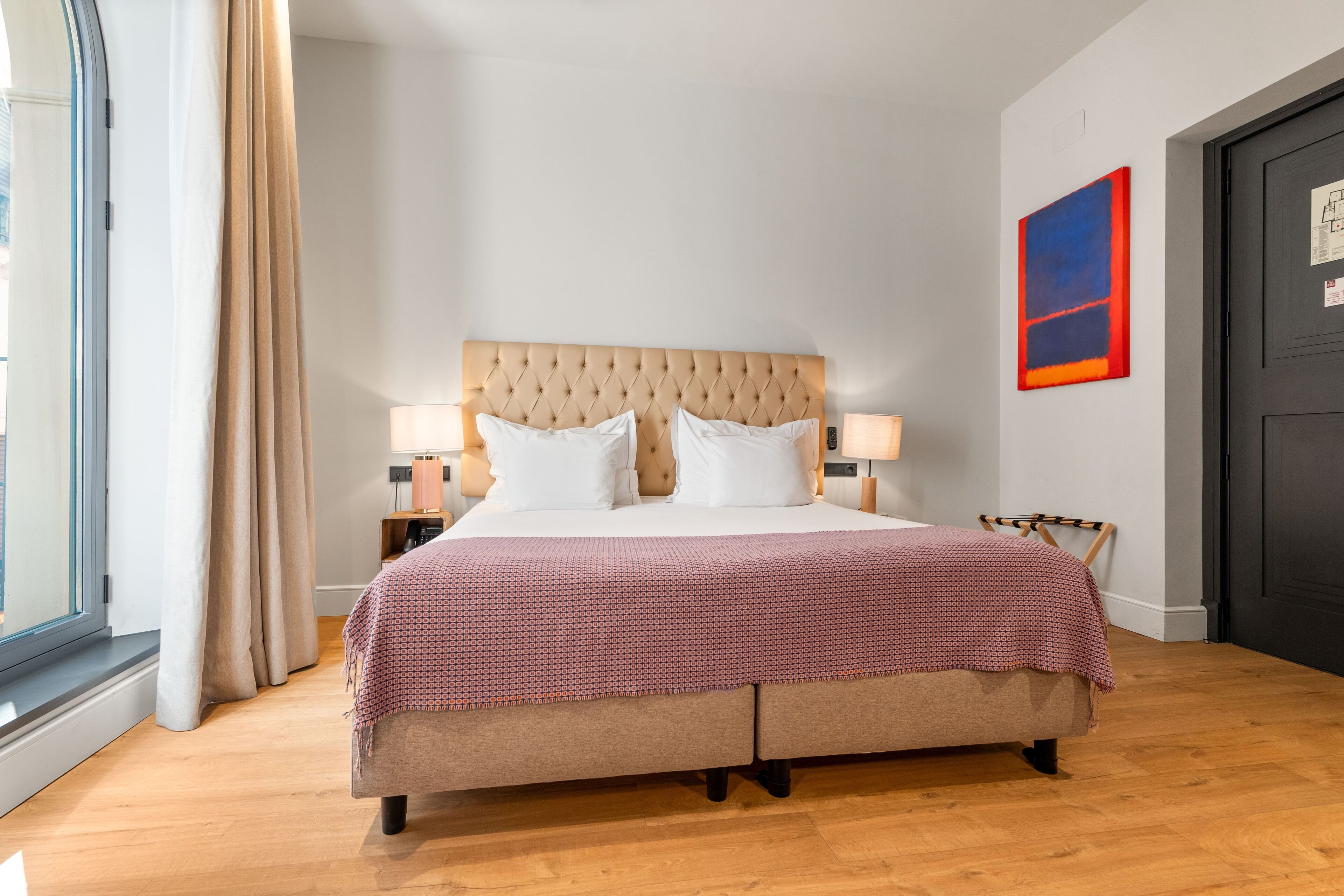 A bedroom with a large bed, two bedside lamps, a window with curtains, and a door with a red and blue painting.