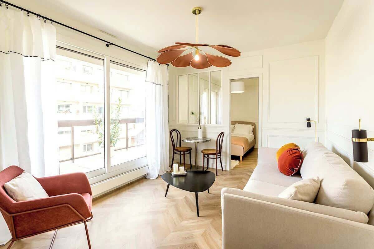 A modern living room with a sofa, armchair, and a dining table with chairs. There is a large window providing natural light and a view of the outside.