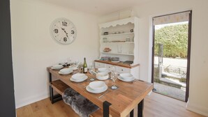 A dining area with a wooden table set for four, a white clock on the wall, and a built-in white shelving unit with kitchen items. - Fawley (Lyme Regis)