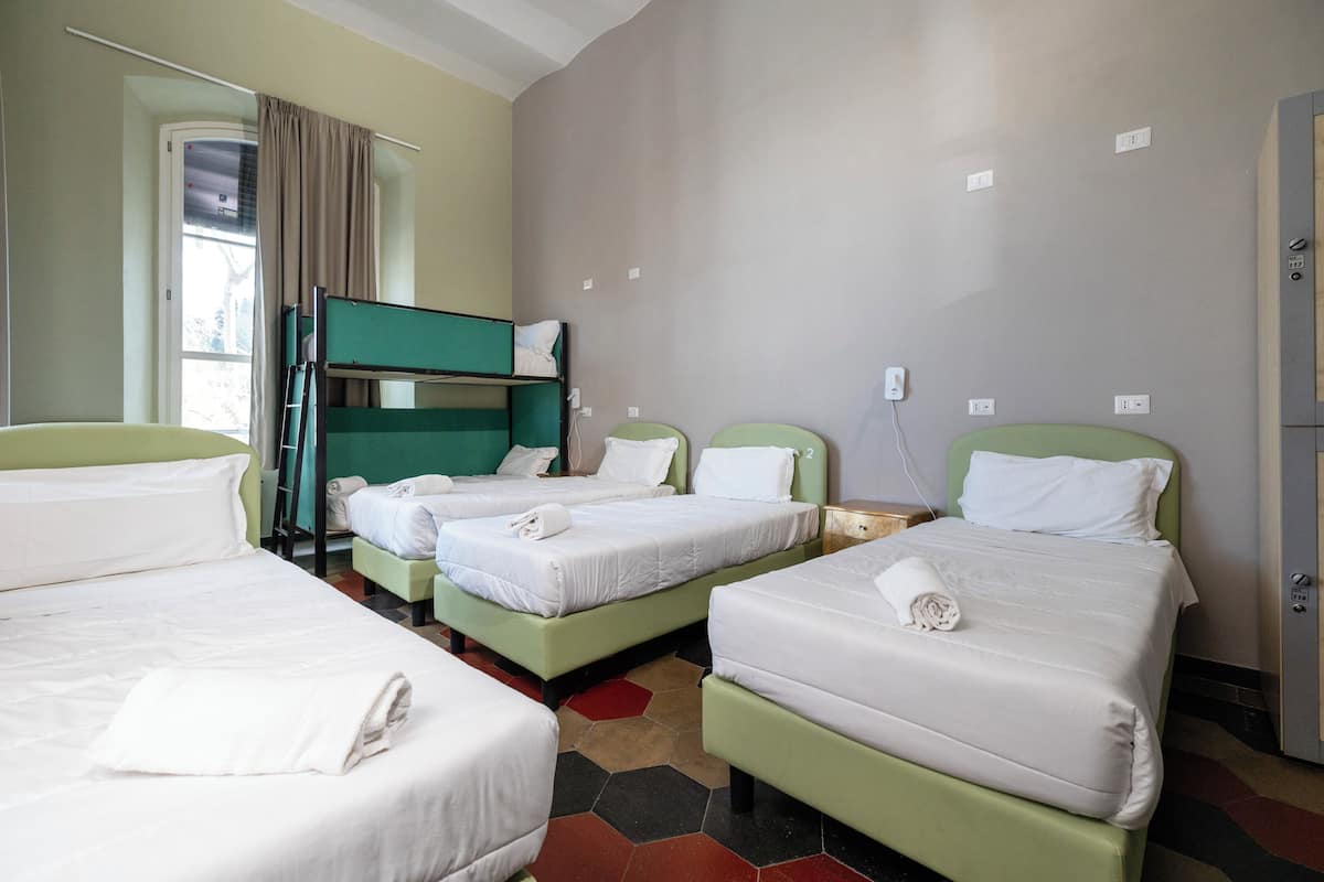 Quarto de hotel com quatro camas alinhadas, uma janela com cortinas e uma porta à direita.