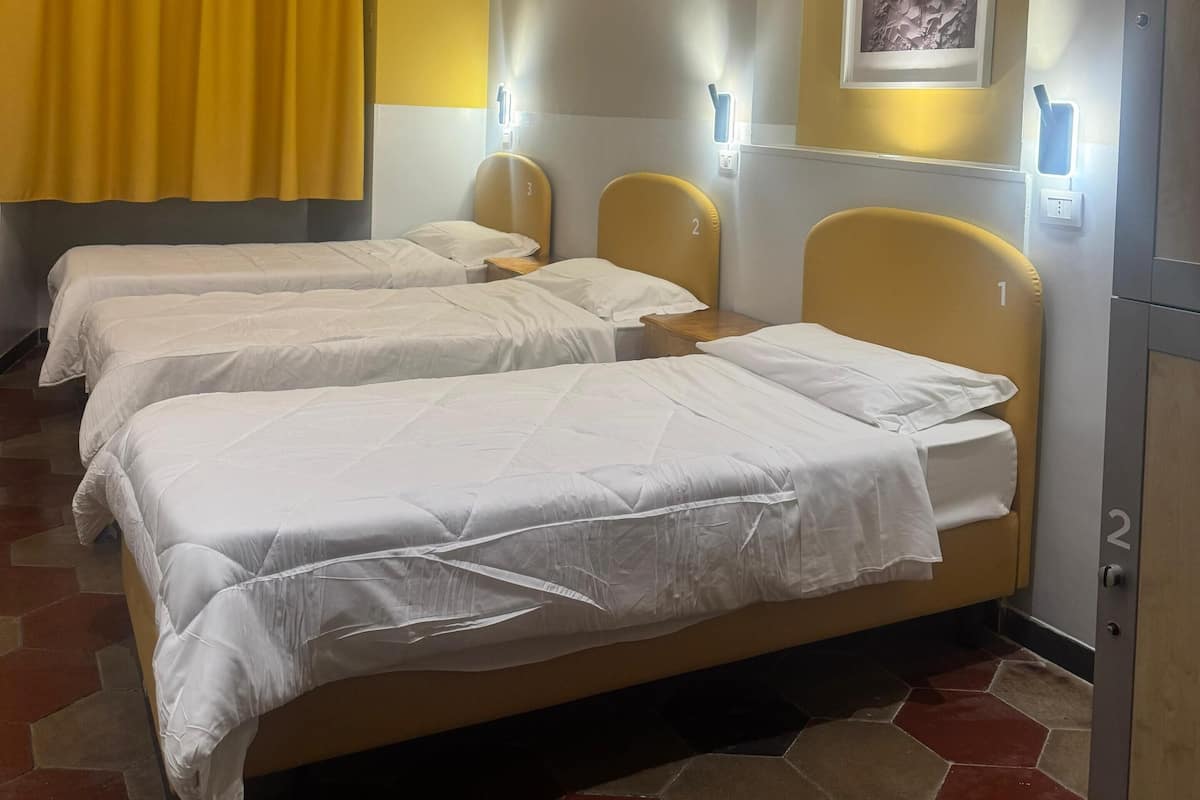 Quarto de hotel com duas camas, uma cortina amarela, um quadro na parede e uma porta com número.