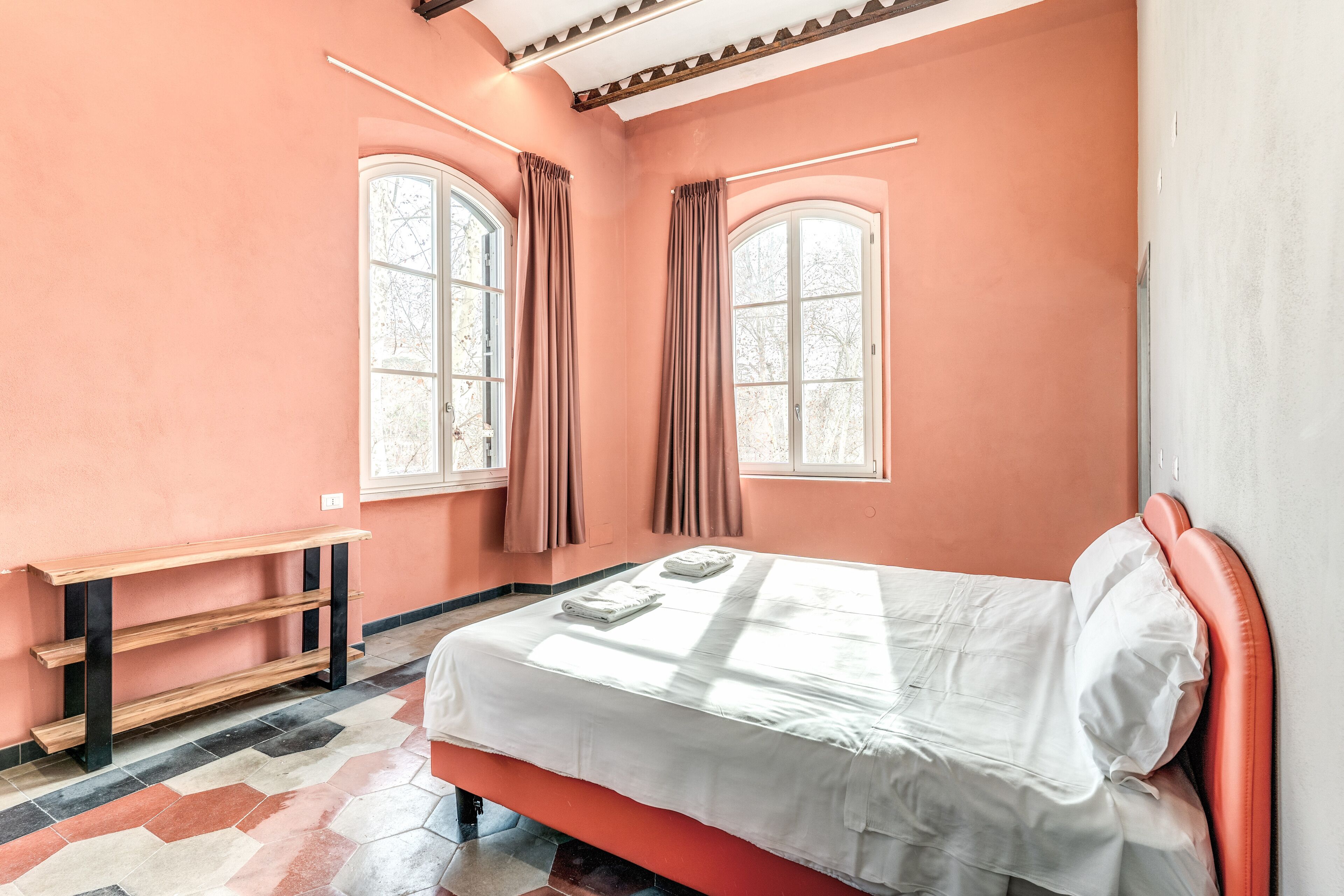 Un dormitorio con una cama, dos ventanas con cortinas, un banco de madera y suelo de baldosas.