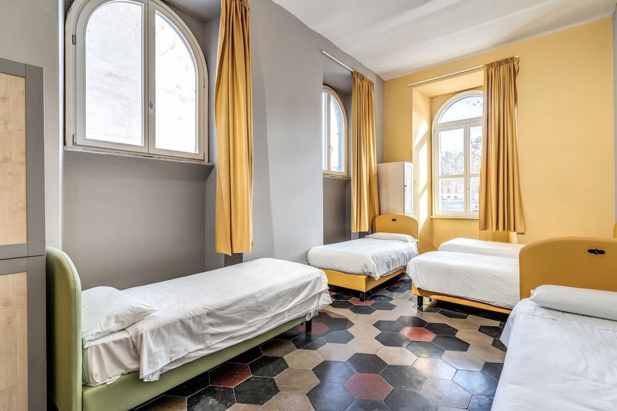 Quarto com três camas, janelas amplas com cortinas amarelas e piso com padrão hexagonal.