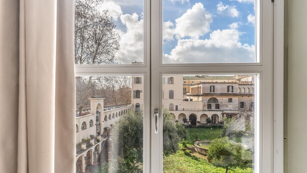 A view through a window of a courtyard with arches, a fountain, and buildings in the background. - Borgo Ripa Urban Travel (Rome)