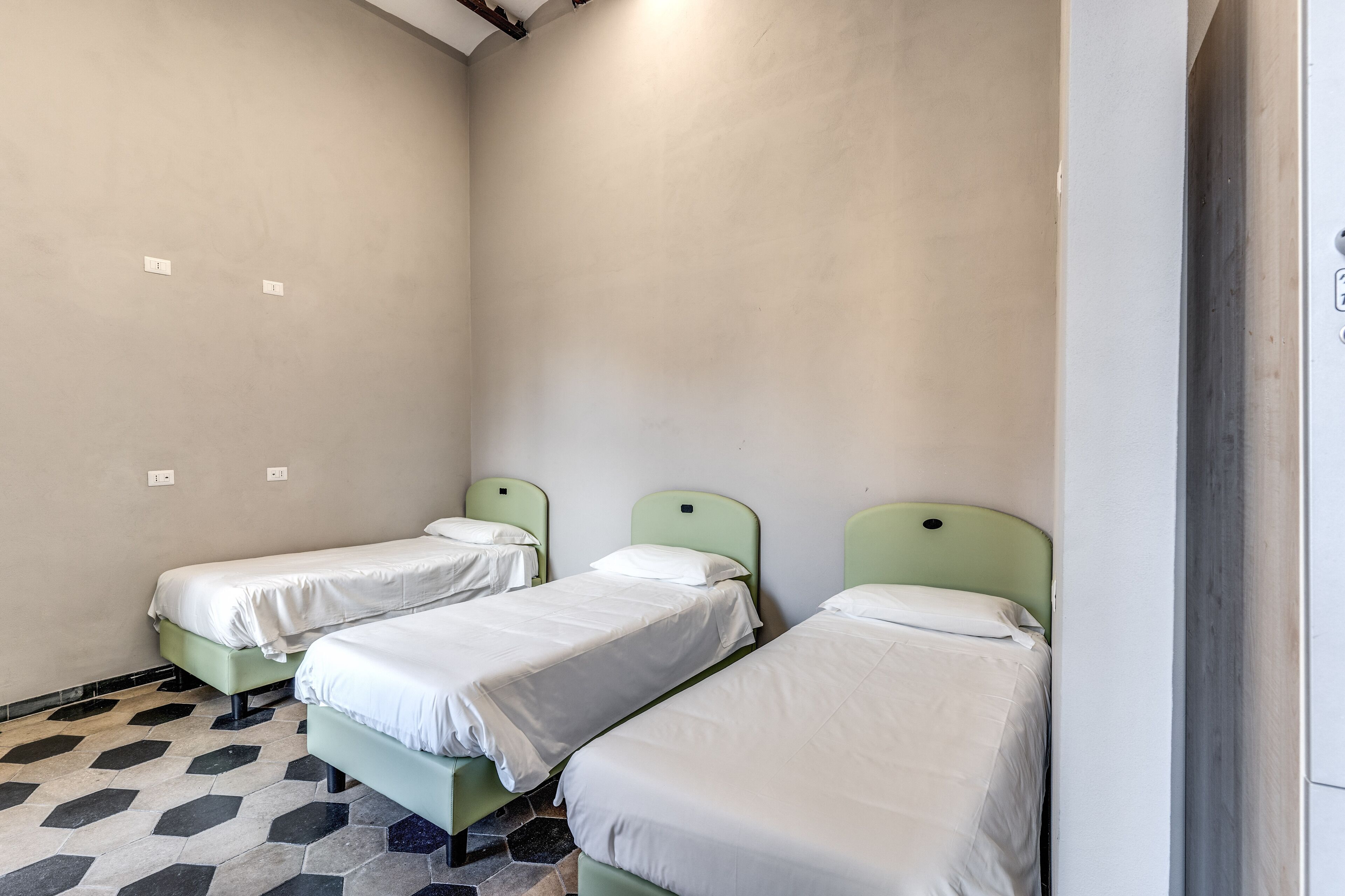 A small, clean room with three single beds, each with a green headboard and white bedding. The floor has a geometric tile pattern.