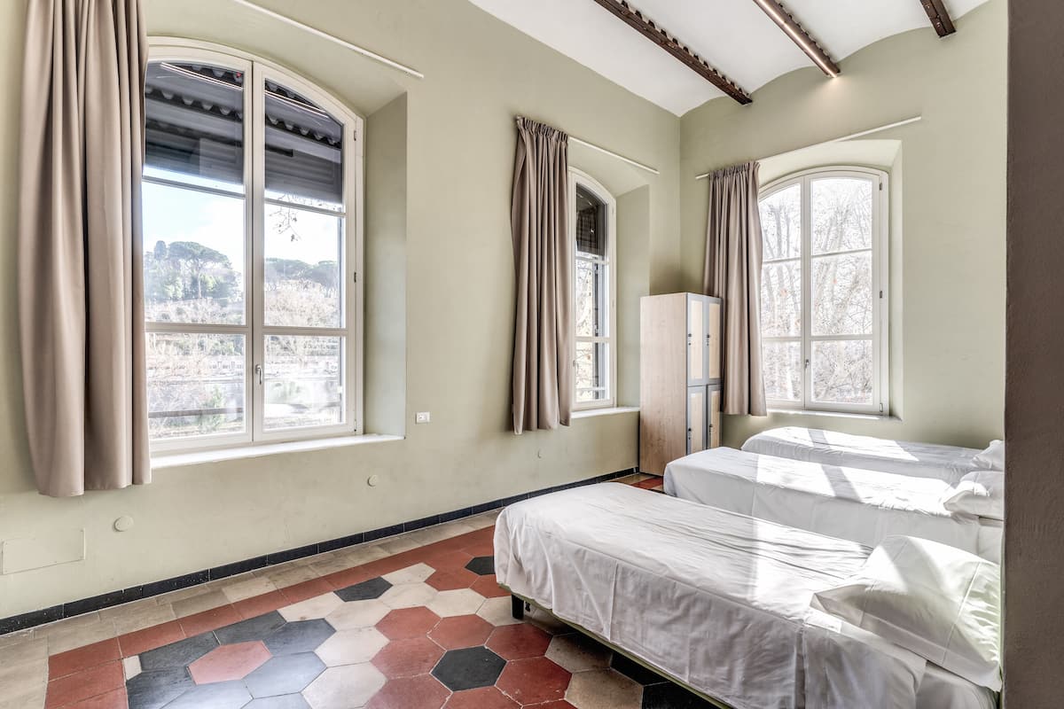 Quarto com duas camas, amplas janelas com cortinas, radiador e piso revestido de azulejos.