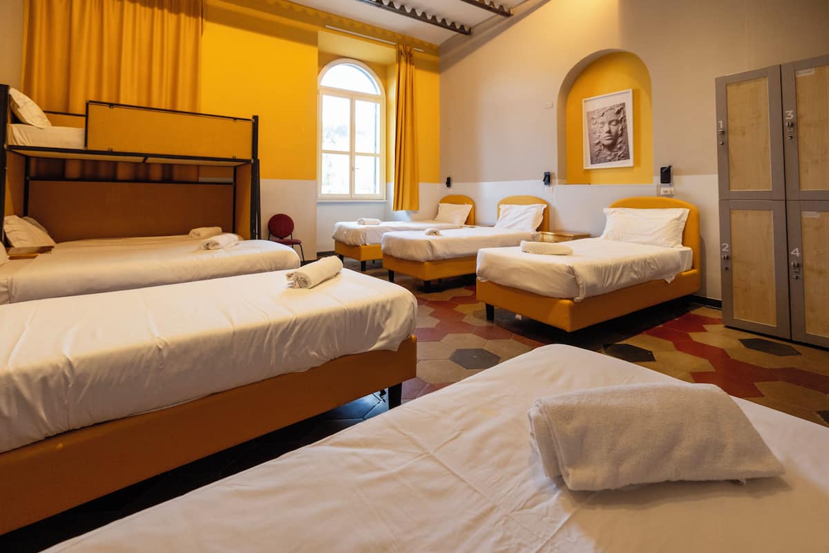 Quarto de hotel com quatro camas, uma janela grande com cortinas, uma obra de arte na parede e um guarda-roupa.