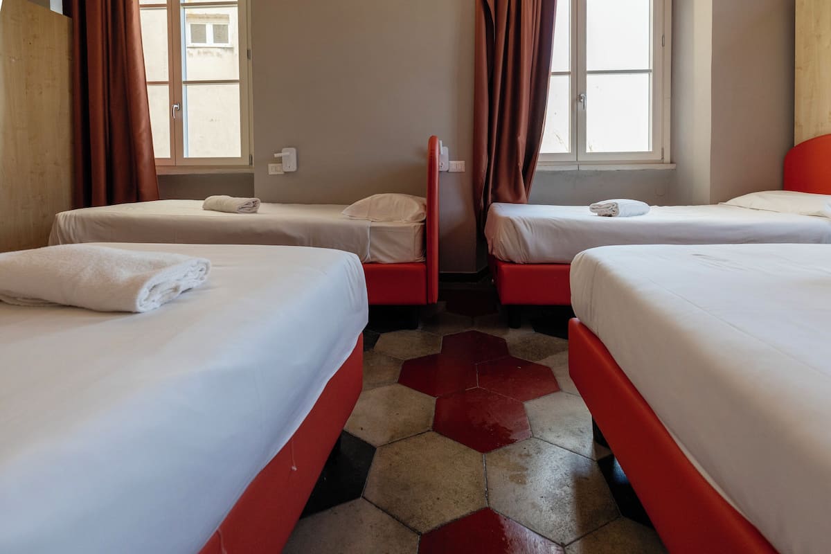 Quarto de hotel com três camas, cada uma com roupa de cama branca e cabeceiras vermelhas. O quarto tem piso de azulejos com padrão hexagonal e uma janela com cortinas marrons.